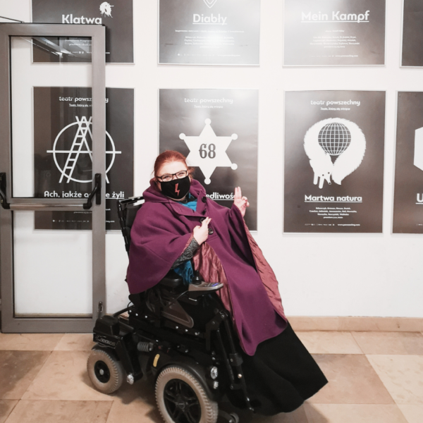 kobieta na elektrycznym wózku inwalidzkim w fioletowym poncho na tle ściany czarno-białych plakatów teatralnych